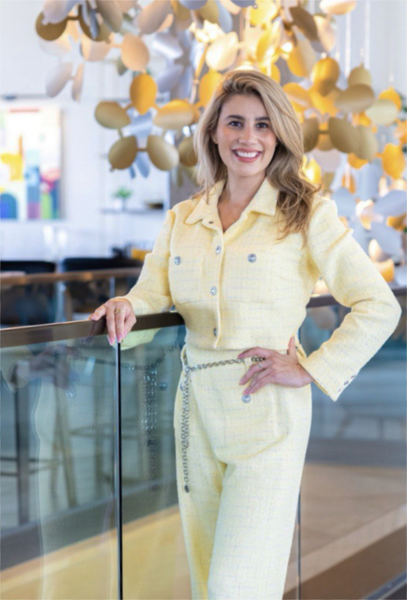 Smiling, elegant blond long hair woman wearing a lemon yellow suit standing next to a glass banister.